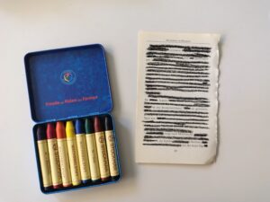 eine Buchseite mit Blackout Poery liegt rechts neben einer blauen Dose mit Wachsmalstiften